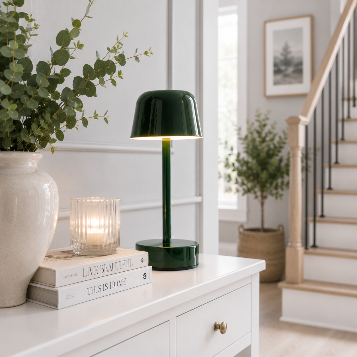 Lampe Nova LED rechargeable vert forêt sur une console de hall d'entrée élégante avec escalier, bougie en verre, livres décoratifs et plante dans un intérieur moderne aux tons greige et bleu poudre.