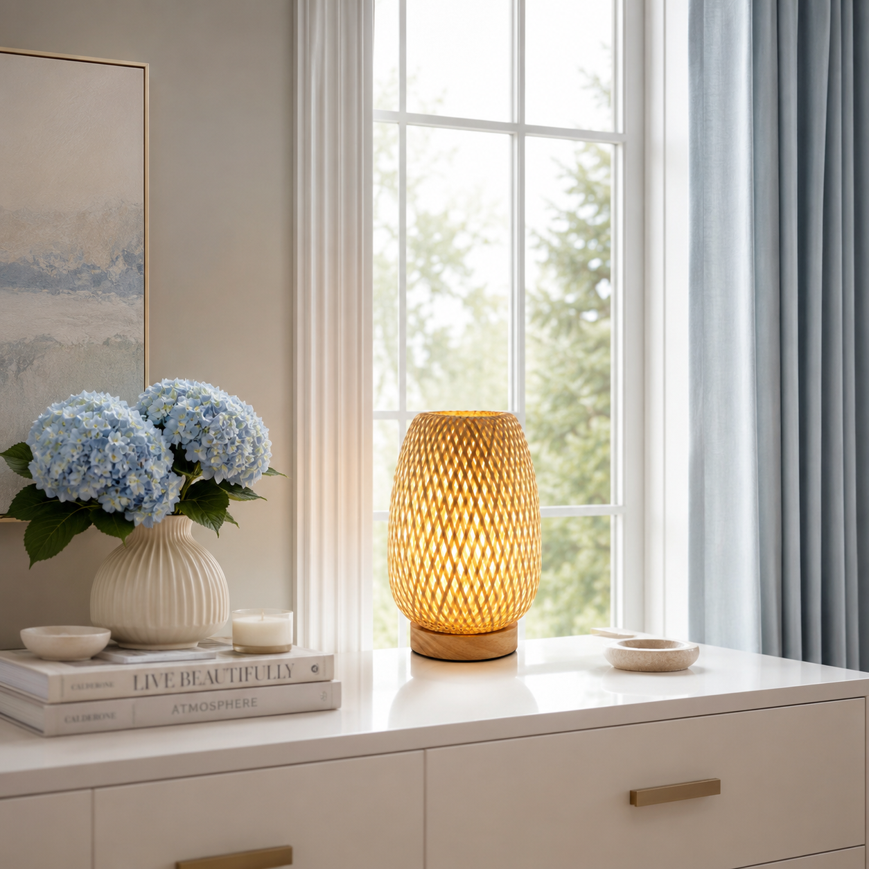 Lampe de table Tressa en bambou tressé à la main diffusant une lumière douce et chaleureuse sur une console blanche devant une grande fenêtre. Son design en bois naturel s'intègre parfaitement à une décoration intérieure moderne avec vase fleuri, lumière naturelle et ambiance élégante aux tons greige, crème et bleu poudre, idéale pour le salon, la chambre ou l'entrée.