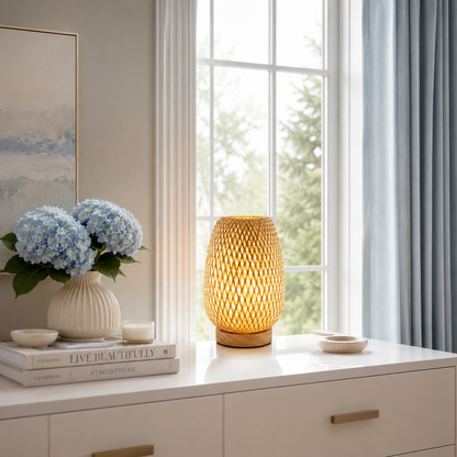 Lampe de table Tressa en bambou tressé à la main diffusant une lumière douce et chaleureuse sur une console blanche devant une grande fenêtre. Son design en bois naturel s'intègre parfaitement à une décoration intérieure moderne avec vase fleuri, lumière naturelle et ambiance élégante aux tons greige, crème et bleu poudre, idéale pour le salon, la chambre ou l'entrée.