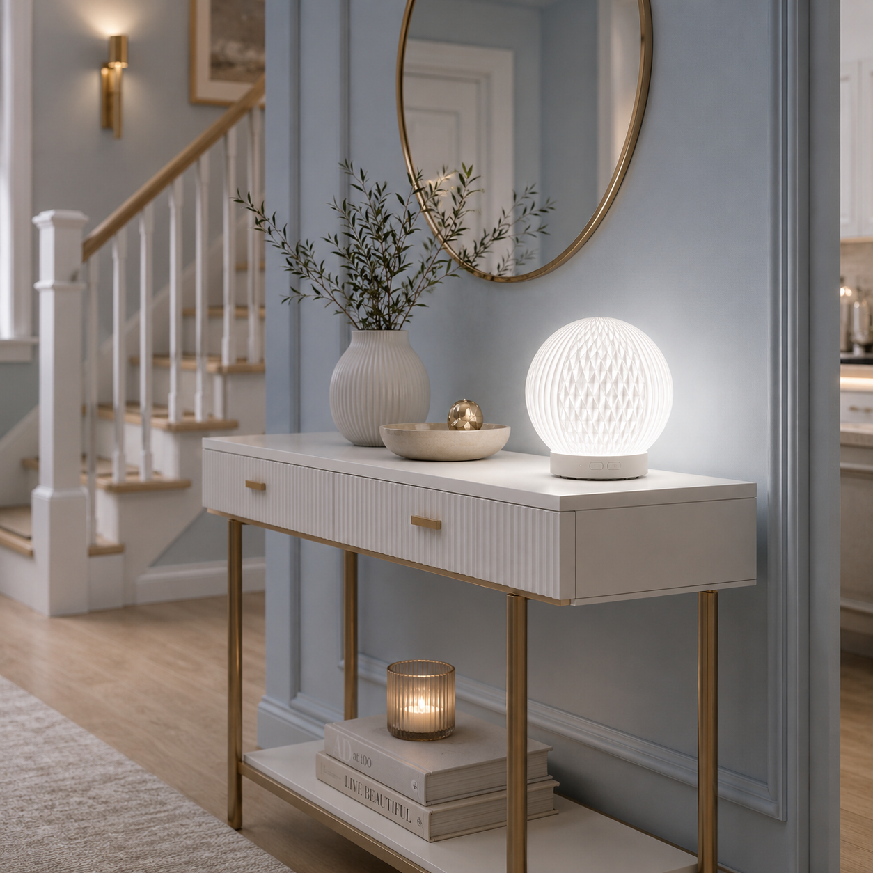 Diffuseur d'huiles essentielles LED Brise allumé sur une console blanche dans un hall d'entrée luxueux aux tons greige et bleu poudre. Décoration raffinée avec miroir rond, escalier, vase, bougie et mobilier haut de gamme créant une ambiance lumineuse, apaisante et chaleureuse.