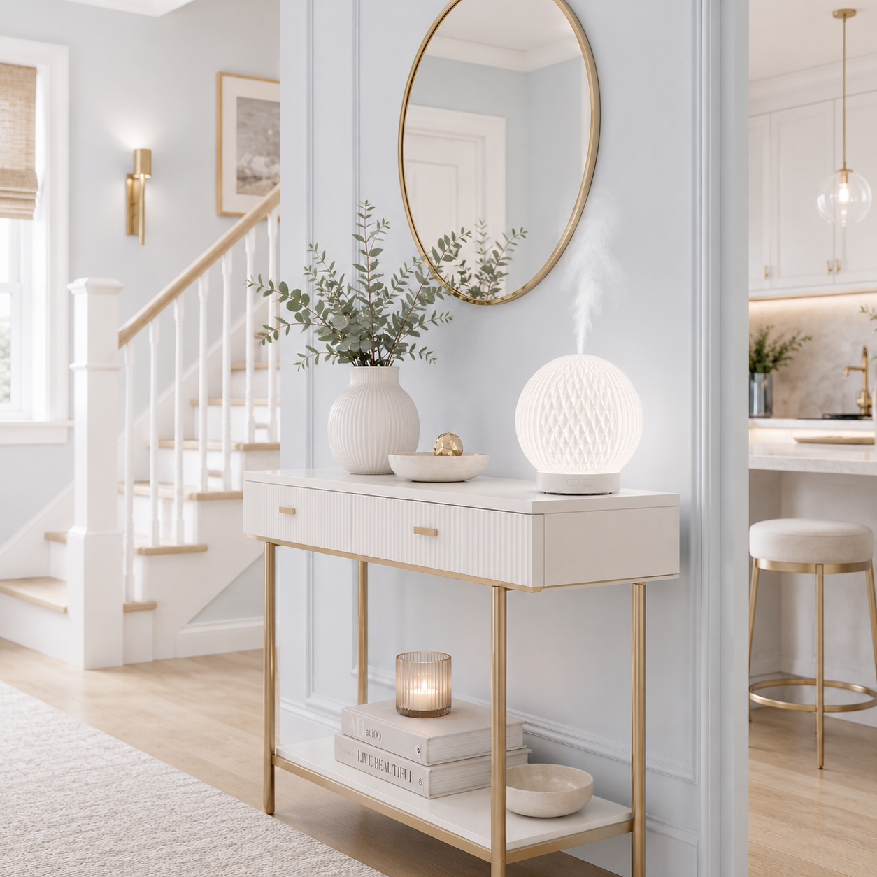 Diffuseur d'huiles essentielles LED Brise diffusant une fine brume sur une console blanche dans un hall d'entrée haut de gamme aux tons greige et bleu poudre. Décoration moderne avec miroir rond, vase et escalier créant une ambiance élégante, relaxante et chaleureuse.