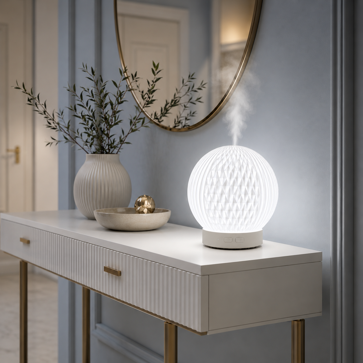 Diffuseur d'huiles essentielles LED Brise diffusant une fine brume sur une console blanche dans un hall d'entrée moderne aux tons greige et bleu poudre. Décoration haut de gamme avec miroir rond, vase, bougie et mobilier blanc créant une ambiance élégante, lumineuse, relaxante et chaleureuse.