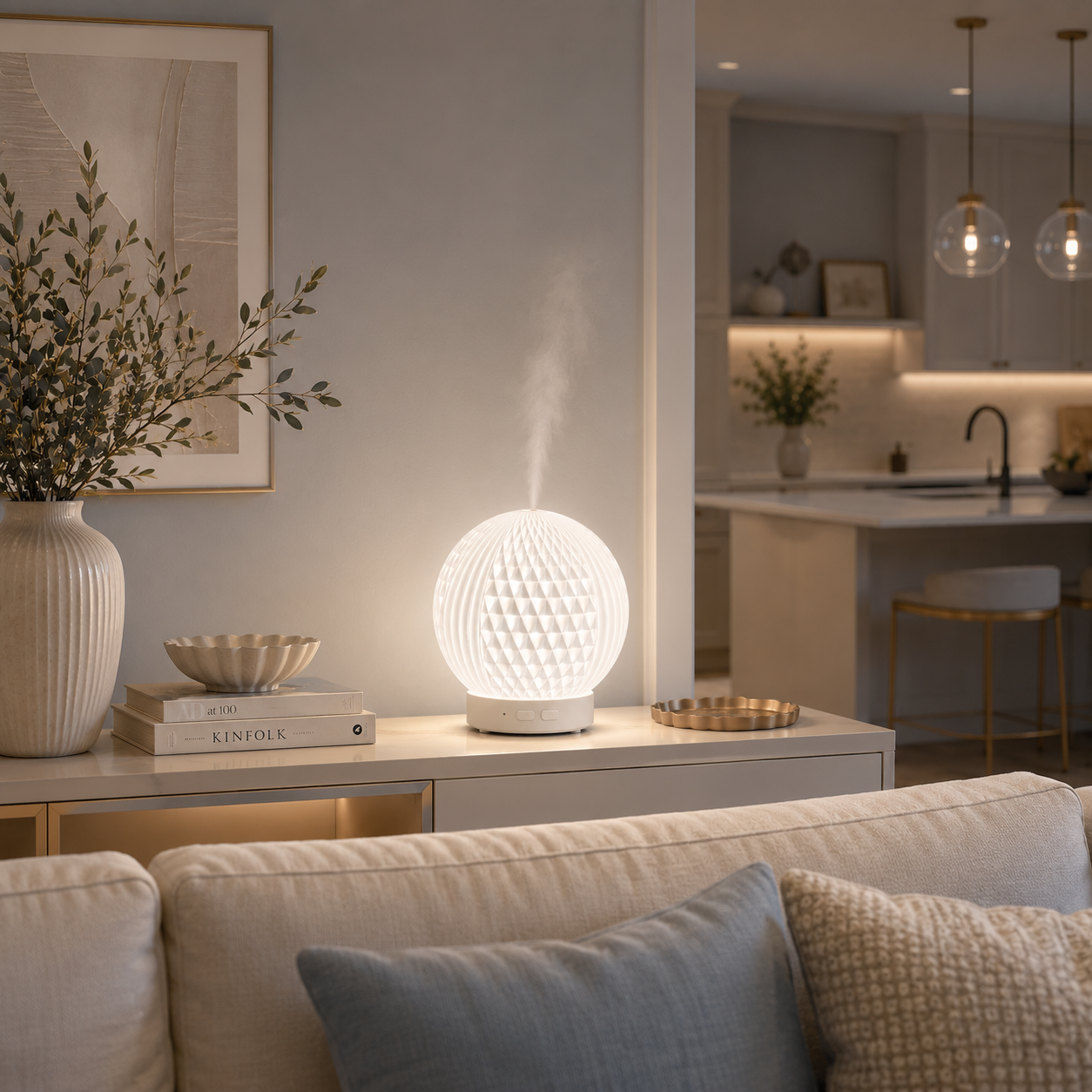 Diffuseur d'huiles essentielles LED Brise diffusant une fine brume sur une console blanche derrière un canapé dans un salon haut de gamme aux tons greige et bleu poudre. Décoration raffinée avec vase, livres, cuisine ouverte et mobilier premium créant une ambiance lumineuse, apaisante et chaleureuse.