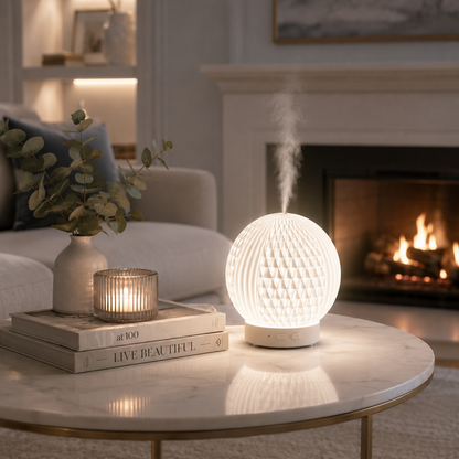 Diffuseur d'huiles essentielles LED Brise diffusant une fine brume sur une table basse en marbre devant un foyer dans un salon haut de gamme aux tons greige et bleu poudre. Décoration élégante avec vase, bougie, livres et lumière chaleureuse créant une ambiance relaxante, apaisante et raffinée.