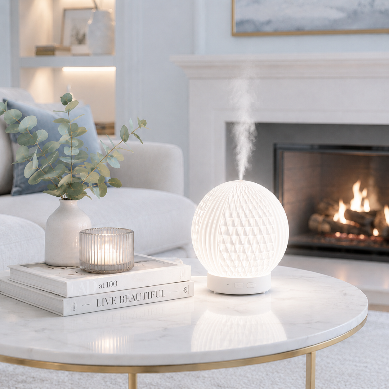 Diffuseur d'huiles essentielles LED Brise diffusant une fine brume sur une table basse en marbre devant un foyer dans un salon haut de gamme aux tons greige et bleu poudre. Décoration élégante avec vase, bougie et livres créant une ambiance chaleureuse, relaxante et raffinée.