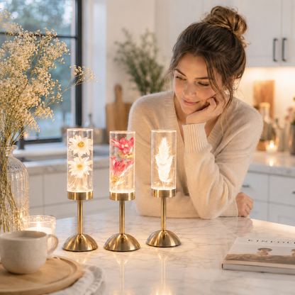 Trois lampes décoratives LED rechargeables en verre avec marguerites blanches, fleurs roses et plume lumineuse, présentées sur un îlot de cuisine en marbre. Une femme admire les lampes dans une cuisine moderne aux tons greige et bleu poudre, créant une ambiance chaleureuse, élégante et apaisante.