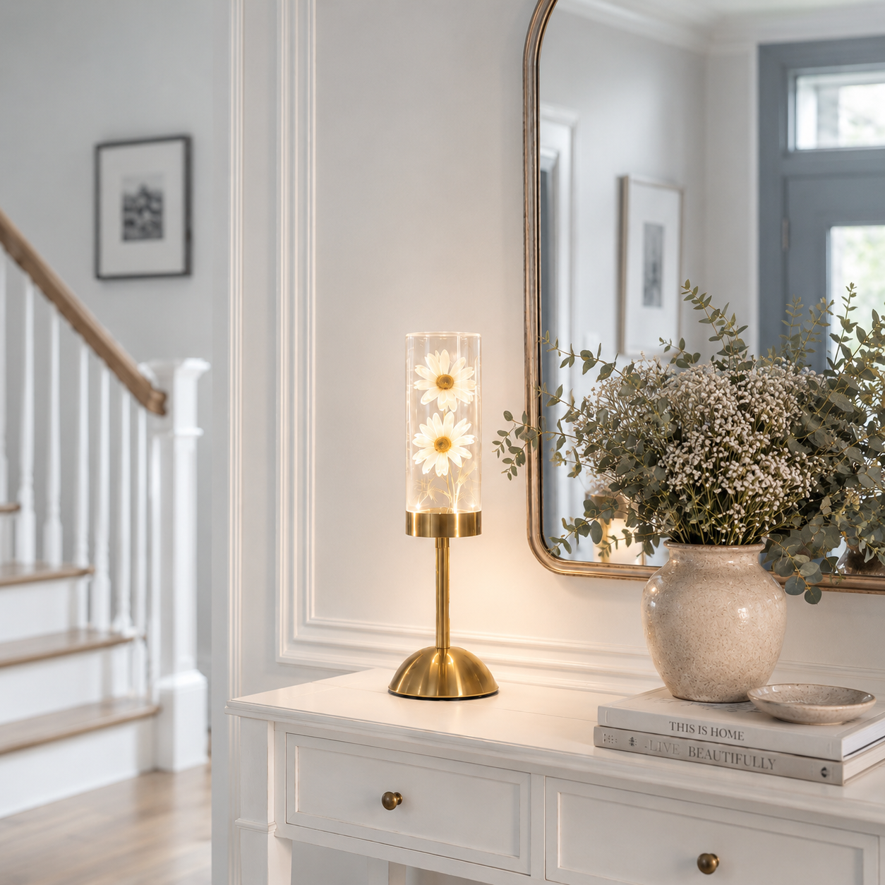 Lampe décorative LED rechargeable avec marguerites blanches illuminées dans un cylindre en verre, mise en valeur sur une console de hall d'entrée moderne avec miroir, vase de fleurs et mobilier blanc aux tons greige et bleu poudre. Une décoration élégante et chaleureuse qui accueille les invités avec une lumière douce et raffinée.