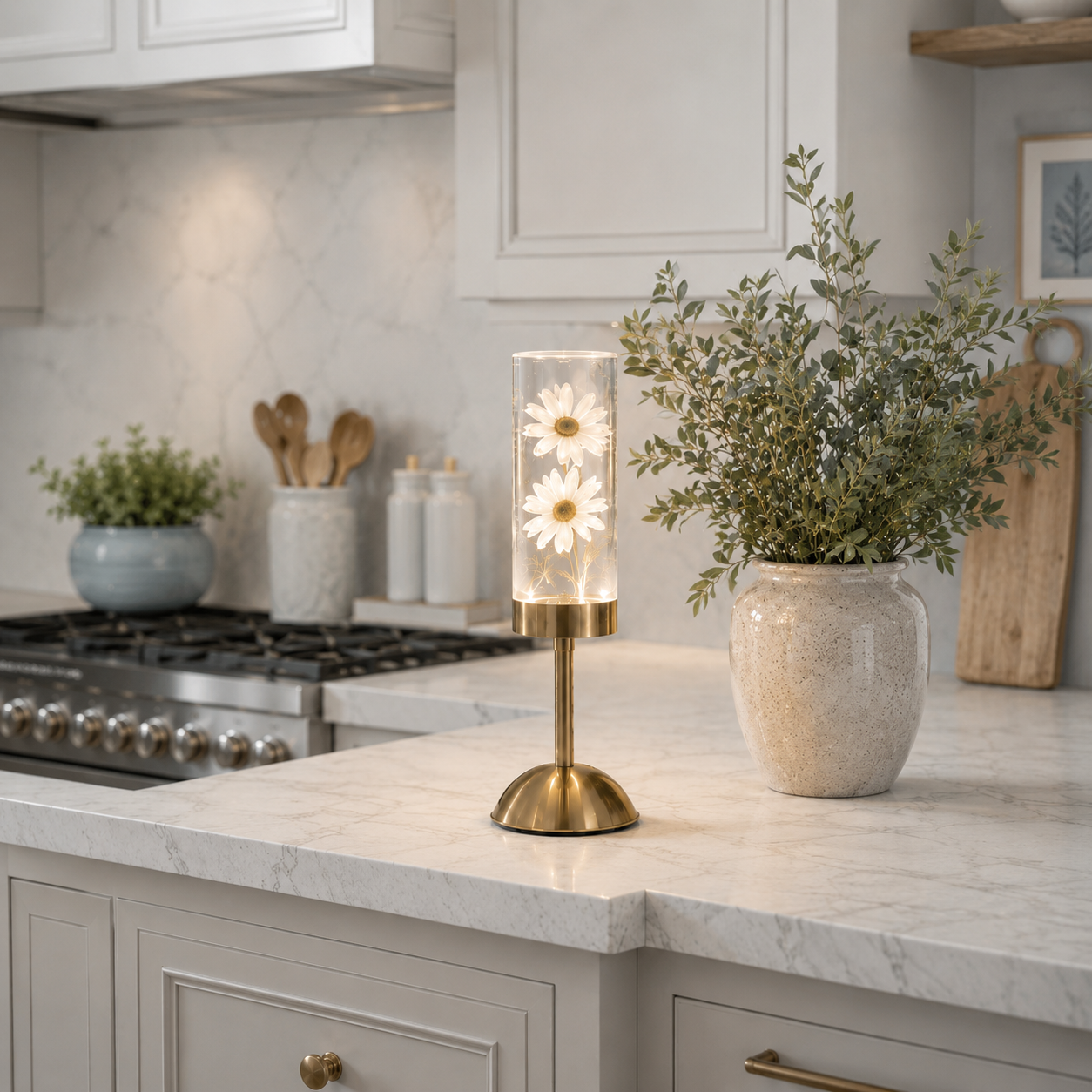 Lampe décorative LED rechargeable avec marguerites blanches illuminées dans un cylindre en verre, posée sur un comptoir de cuisine en quartz dans un intérieur moderne aux tons greige et bleu poudre. Entourée d'un grand vase de verdure, d'ustensiles en bois et d'une cuisine élégante, cette lampe diffuse une lumière douce et chaleureuse qui apporte une ambiance raffinée, apaisante et accueillante.