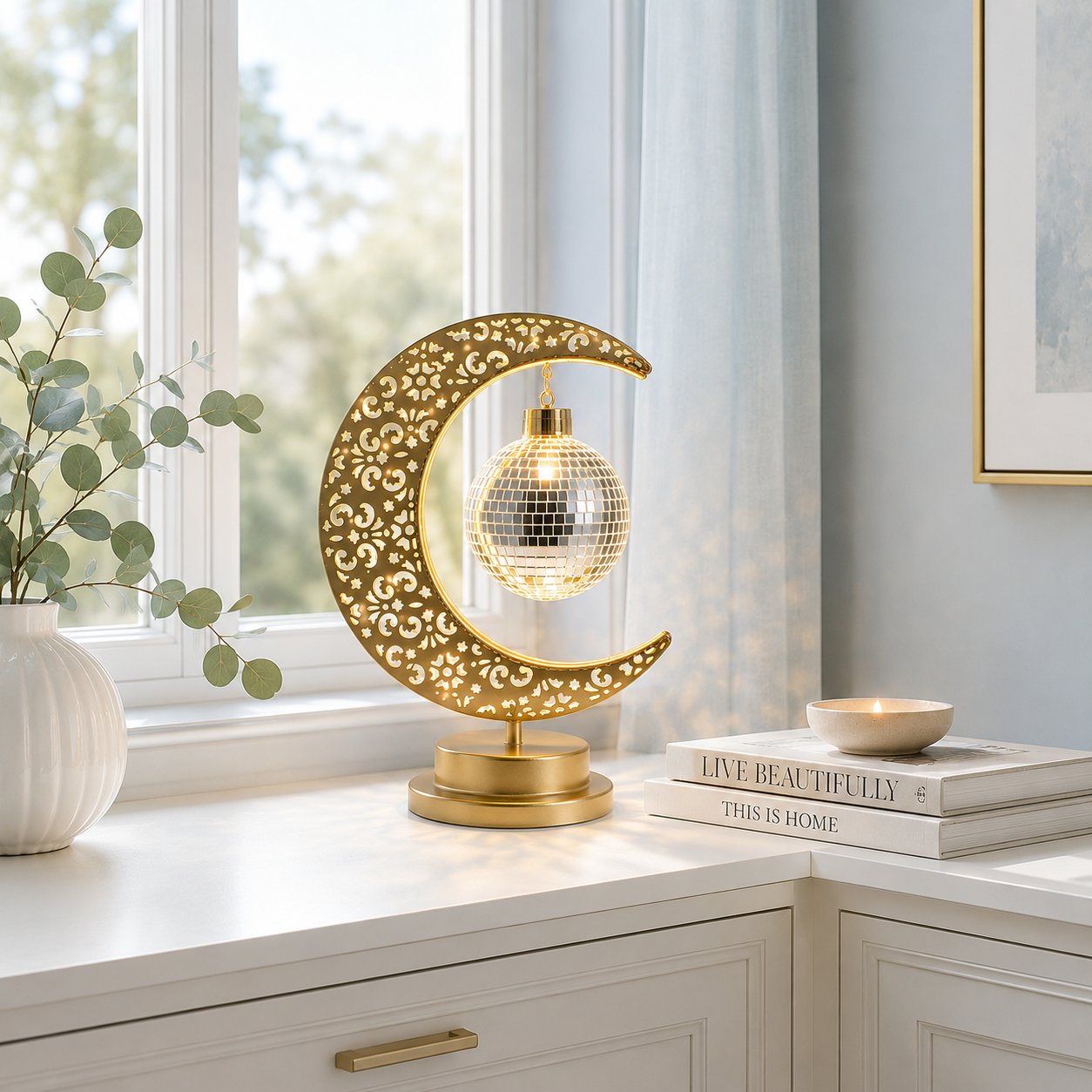 Lampe Lune Enchantée Boule Miroir dorée allumée sur une console blanche près d'une fenêtre, éclairage LED décoratif créant une ambiance douce et chaleureuse dans un intérieur élégant.
