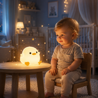 Veilleuse canard en silicone diffusant une lumière douce sur une petite table en bois, devant un bébé assis sur une chaise dans une chambre aux teintes bleu poudre. De délicates étincelles dorées, un lit de bébé et un éclairage chaleureux créent une ambiance rassurante et féérique pour le coucher.