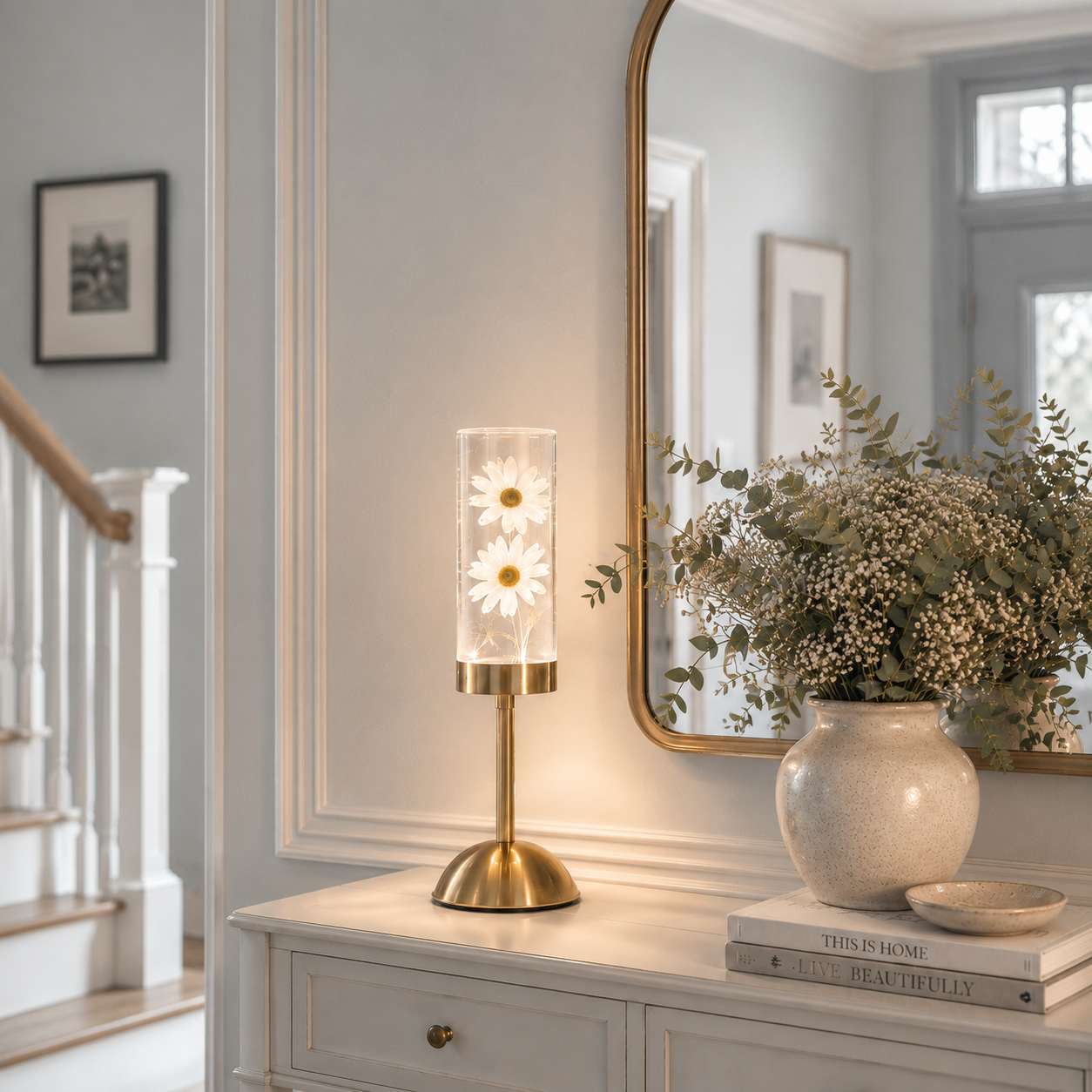 Lampe décorative LED rechargeable avec marguerites blanches illuminées dans un cylindre en verre, mise en valeur sur une console de hall d'entrée élégante près d'un escalier. Entourée d'un grand miroir, d'un vase de fleurs séchées et d'une décoration aux tons greige et bleu poudre, cette lampe diffuse une lumière chaleureuse qui crée une ambiance raffinée et accueillante.
