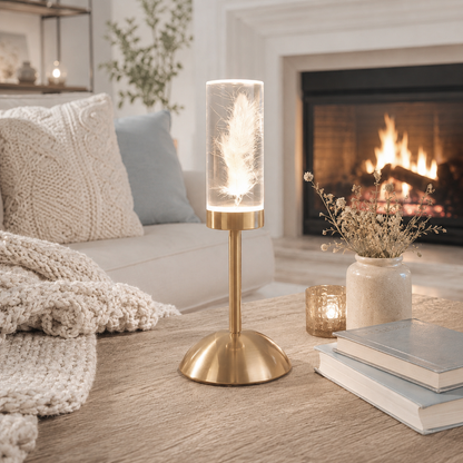 Lampe décorative LED rechargeable avec plume lumineuse dans un cylindre en verre, posée sur une table basse en bois devant un foyer. Entourée de coussins en tricot, de livres décoratifs, d'un vase de fleurs séchées et d'une ambiance aux tons greige et bleu poudre, cette lampe diffuse une lumière douce et chaleureuse pour créer un salon élégant, apaisant et réconfortant.